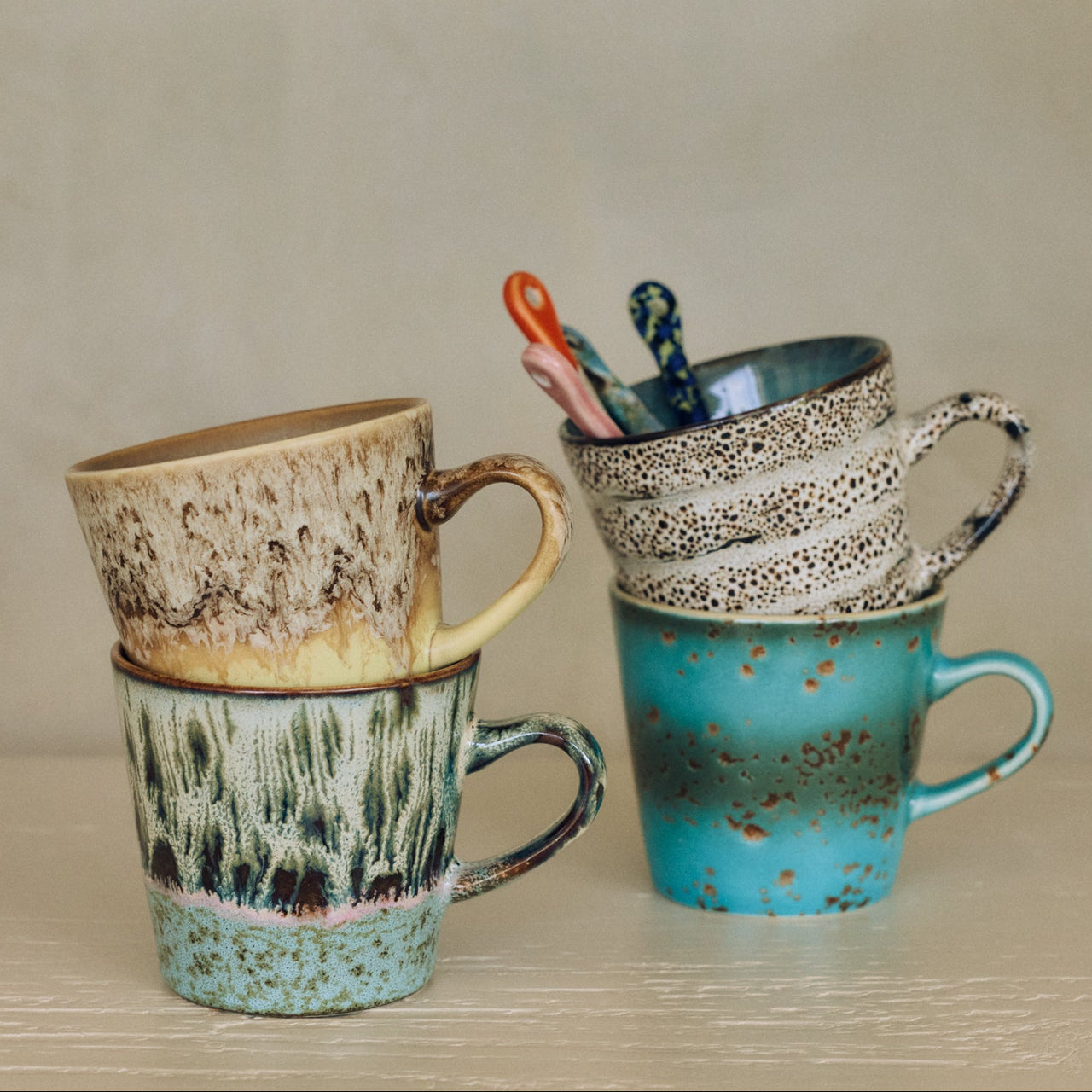 Three handmade ceramic mugs stacked on a light wooden surface against a neutral beige background. The mug on the left is stacked on top of a green and brown patterned mug, both in a warm beige-brown glaze.