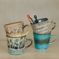 Thumbnail for Three handmade ceramic mugs stacked on a light wooden surface against a neutral beige background. The mug on the left is stacked on top of a green and brown patterned mug, both in a warm beige-brown glaze.
