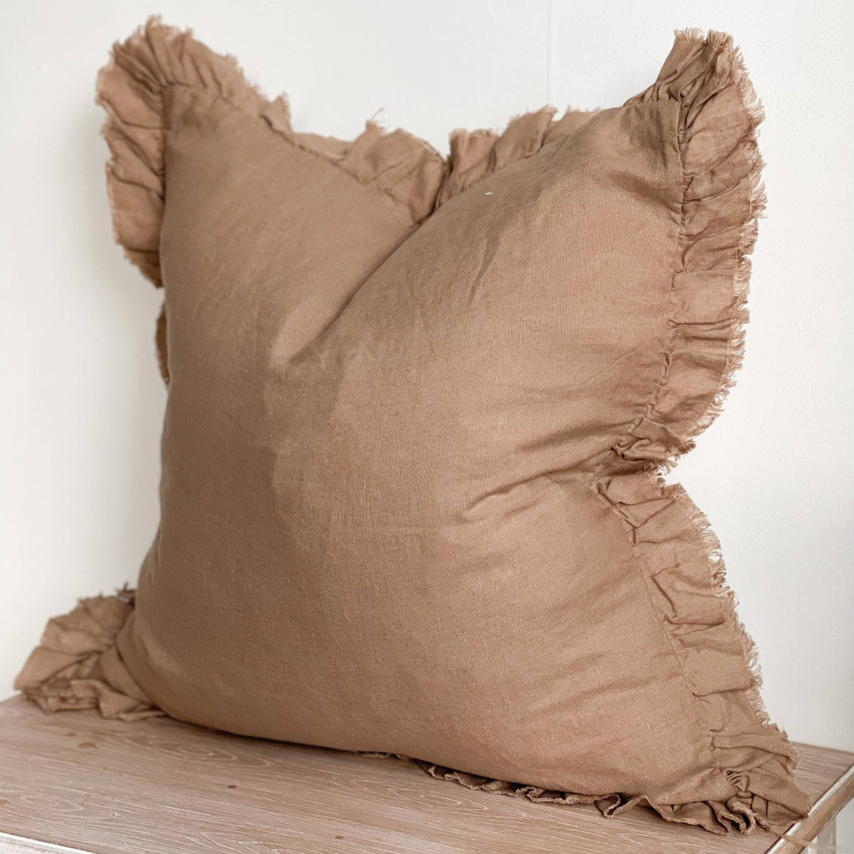 Brown linen pillow with ruffled edges on a wooden surface against a white wall 