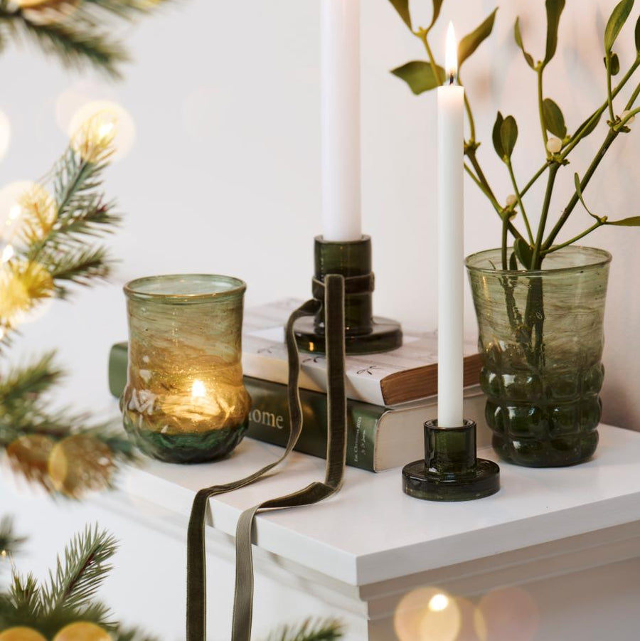 Decorative setup with candles, green glassware, and mistletoe on a white surface with blurred lights in the background.